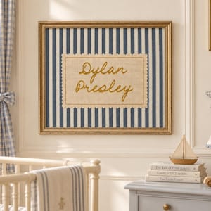 May include: A rectangular wall art piece in a gold frame featuring a navy blue and white vertical striped background. Centered is a cream-coloured fabric patch with scalloped edges, embroidered with the name Dylan Presley in gold cursive script.