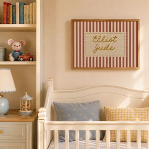 May include: A rectangular wall art piece in a wooden frame featuring a red and white vertical striped background with a central cream-colored scalloped label containing the gold cursive text Elliot Jude, displayed above a white crib.