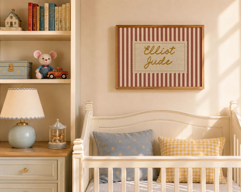 May include: A rectangular wall art piece in a wooden frame featuring a red and white vertical striped background with a central cream-colored scalloped label containing the gold cursive text Elliot Jude, displayed above a white crib.