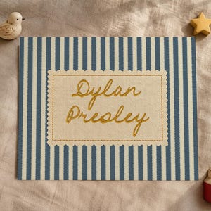 May include: A rectangular fabric sign featuring blue and white vertical stripes with a central cream-colored scalloped patch embroidered with the name Dylan Presley in gold thread, accompanied by a small bird and star figurine.