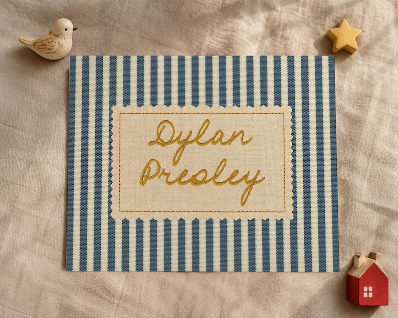May include: A rectangular fabric sign featuring blue and white vertical stripes with a central cream-colored scalloped patch embroidered with the name Dylan Presley in gold thread, accompanied by a small bird and star figurine.