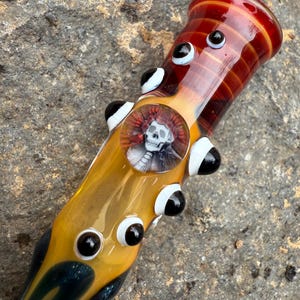 May include: A glass pipe with a red and yellow design. The pipe features a skull image and several black and white eye-shaped embellishments. The pipe is on a textured gray surface.