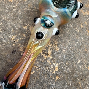 May include: A decorative glass pipe with a rounded bowl and a long, tapered stem. The pipe features an eagle emblem and is decorated with black and white accents. The glass has a mix of colours, including yellow, orange, and blue.