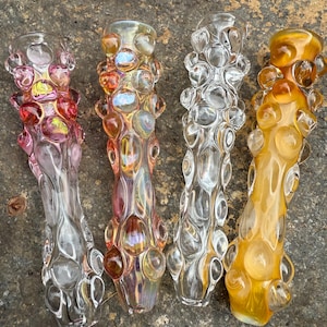 May include: Four glass smoking pipes with a textured, bubble-like design. The pipes vary in color, including pink, iridescent, clear, and amber. Each pipe has a cylindrical shape with a flared mouthpiece.