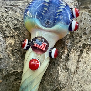 May include: A hand-blown glass pipe with a blue and white patterned bowl. The pipe features red, white, and blue accents, including a small glass marble with an American flag design. The stem has a swirling, translucent design.