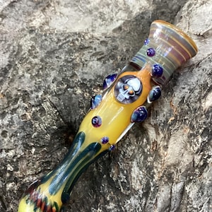 Könnte beinhalten: Eine gelbe Glaspfeife mit Totenkopf-Design. Die Pfeife hat einen zylindrischen Körper mit farbigen Akzenten, darunter blaue und violette Glasperlen. Das Ende der Schale hat ein Streifenmuster, und das Mundstück hat ein detailliertes, strukturiertes Design.