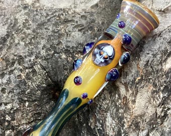 May include: A yellow glass pipe with a skull motif. The pipe has a cylindrical form with colourful accents, including blue and purple glass beads. The bowl end has a striped pattern, and the mouthpiece has a detailed, textured design.
