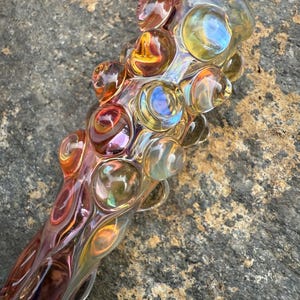 Könnte beinhalten: Eine farbenfrohe Glaspfeife mit einer Reihe von abgerundeten, blasenartigen Vorsprüngen. Das Glas weist eine Mischung aus Orange-, Gelb- und Lilatönen mit irisierenden Eigenschaften auf. Die Pfeife ist mit einer gebogenen Form gestaltet.