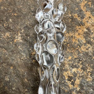 Puede incluir: Un jarrón decorativo de vidrio transparente con un diseño texturizado en forma de burbujas. El jarrón tiene una parte superior ancha y un cuerpo largo y delgado. El vidrio transparente permite el paso de la luz. El jarrón está sobre una superficie marrón texturizada.