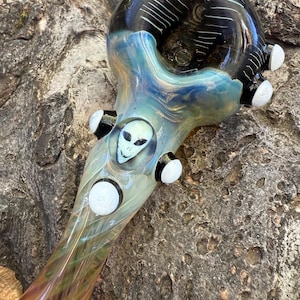 May include: A hand-blown glass pipe with an alien face design. The pipe features a black and white bowl, a blue and green body, and white accents. The alien face is in the center of the pipe.