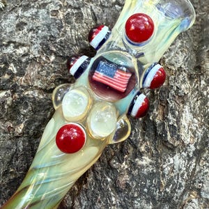 May include: A clear glass pipe with a patriotic theme. The pipe features red, white, and blue accents, including small red spheres and an embedded American flag. The glass has a swirling, iridescent finish.