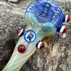 May include: A glass pipe with a blue and white bowl, transitioning to a yellow and green stem. The pipe features red, white, and blue accents, and a small Pabst Blue Ribbon logo.