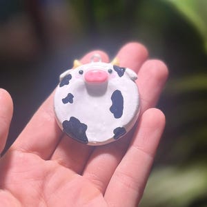 Könnte beinhalten: Handgefertigter, runder Kuh-Ornament. Das Ornament ist weiß mit schwarzen Flecken, einer rosa Nase und kleinen gelben Hörnern. Es hat eine kleine Schlaufe oben zum Aufhängen. Der Hintergrund ist unscharf.