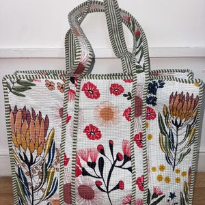 May include: A floral patterned tote bag. The bag has a white background with red, orange, and blue flowers. The handles and edges are trimmed with green and white striped fabric. The bag is designed for carrying items, and is made of quilted fabric.