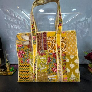 May include: A patchwork tote bag featuring floral and geometric patterns in yellow, green, and red hues. The bag has a rectangular shape with a striped handle and a cream trim. The bag is made of fabric.