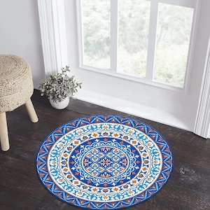 May include: A round blue rug with an intricate pattern of white, orange, and blue geometric designs. The rug is placed on a dark wood floor near a window and a woven stool. The rug's design features a central medallion and a border.