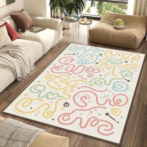 May include: A rectangular area rug with a cream-colored background and a colorful, whimsical design. The rug features a maze-like pattern with lines in pink, yellow, blue, and green, with arrows and other shapes. The rug is placed on a hardwood floor.
