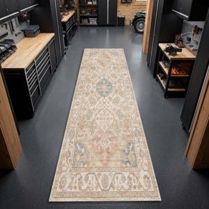 May include: A long rectangular runner rug featuring a faded, intricate pattern in shades of beige, muted orange, and slate blue. The rug is centered on a dark gray floor in a workshop setting with black metal cabinets and wooden workbenches.