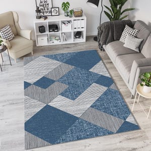 Könnte beinhalten: Ein rechteckiger Teppich mit einem geometrischen Muster in Blau-, Grau- und Weißtönen. Das Design weist verschiedene Formen und Texturen auf und erzeugt einen modernen und stilvollen Look. Der Teppich liegt auf einem hellen Holzboden.
