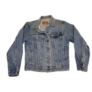 May include: A light blue denim jacket with a button-up front, two chest pockets, and two side pockets. The jacket has a classic collar and long sleeves. A small red tag is visible on the chest pocket.