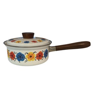 May include: A white enamel saucepan with a wooden handle and lid. The pan is decorated with a floral pattern in red, blue, and yellow. The handle is approximately 8 inches long.