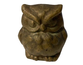 May include: A small, solid bronze-toned owl figurine with a weathered, textured surface. The owl features prominent, defined eyebrows, rounded ears, and a compact, rounded body shape with visible feather detailing across the chest and wings.