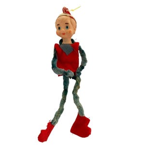May include: A vintage Christmas ornament in the shape of a doll. The doll has a painted face, a red and gray outfit, and red boots. The arms and legs are made of a fuzzy, gray material. The doll is wearing a red and white striped hat.