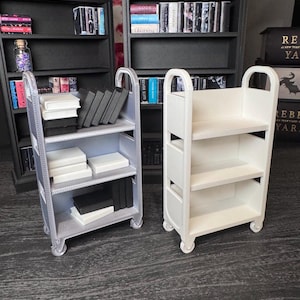 May include: Miniature dollhouse furniture set featuring two rolling carts and two bookshelves. One cart is silver, the other is white. The bookshelves are black. The shelves are filled with miniature books and decorative items.