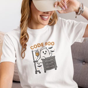 Może przedstawiać: Biała koszulka z napisem "CODE BOO" i postacią z kreskówki ducha w mundurze pielęgniarki, z nietoperzami i medycznym kroplówką. Duch jest na wózku szpitalnym. Modelka ma na sobie beżową czapkę z daszkiem.