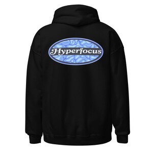 Puede incluir: Una sudadera con capucha negra que presenta un gráfico en la parte posterior. El gráfico es un óvalo azul que contiene un patrón azul claro similar a un cerebro y la palabra Hyperfocus escrita en una fuente serif blanca en el centro.