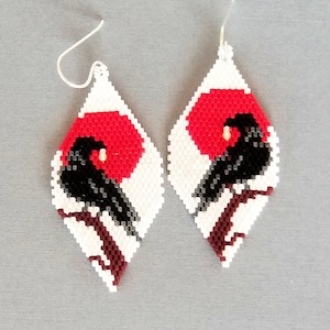 May include: A pair of white teardrop-shaped earrings with a black bird and red sun design. The earrings are made of beads and have a silver hook.
