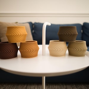 May include: A collection of six small, cylindrical planters with a textured, chevron-patterned exterior. The planters are arranged in various earth tones, including shades of dark brown, mustard yellow, beige, and tan, resting on a white table.