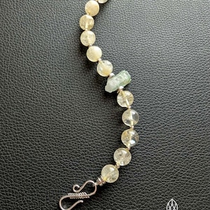 May include: A beaded bracelet featuring pale yellow and clear crystal beads, with a small green crystal accent. The bracelet has a silver clasp and a silver loop at the other end. The bracelet is on a black surface.