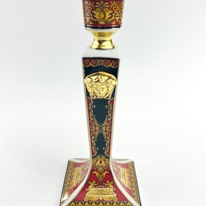 May include: A decorative ceramic candlestick holder with a gold, red, and dark teal color scheme. The holder features ornate gold detailing, a Medusa head emblem, and a square base. The top is designed to hold a single candle.