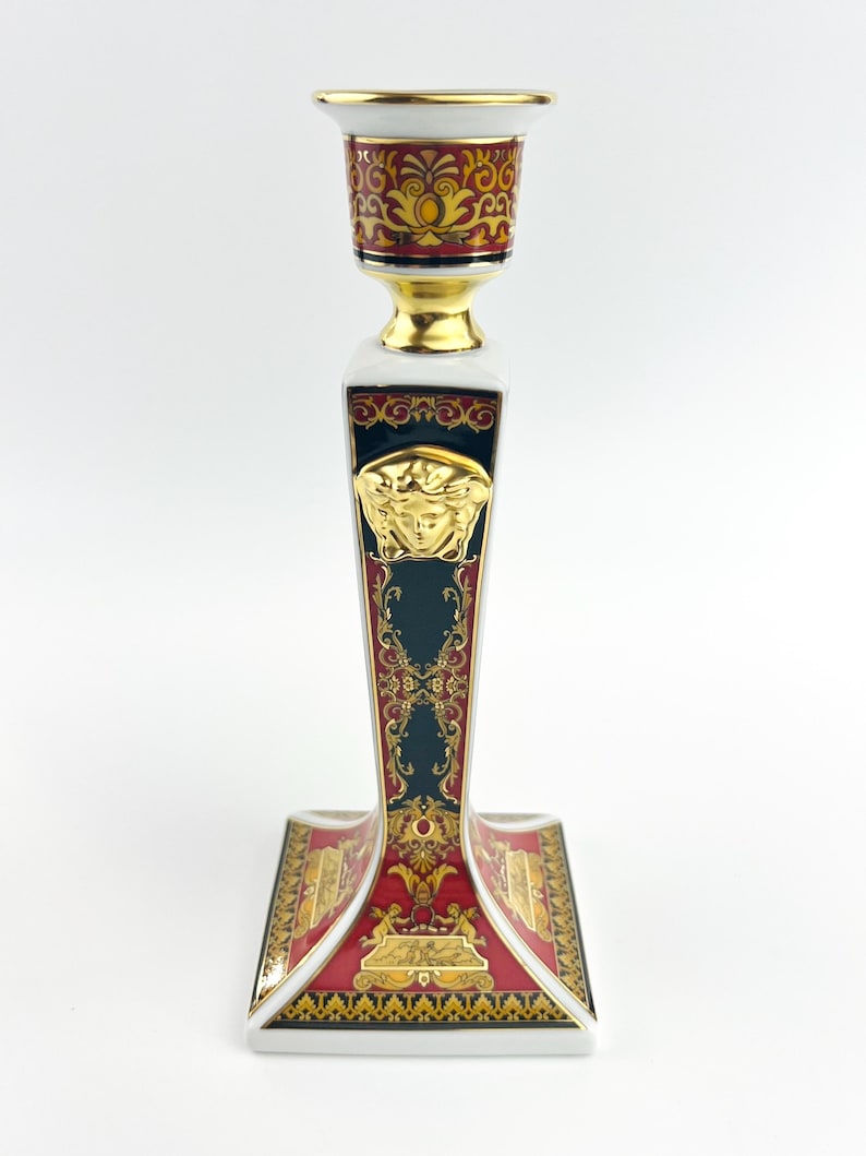 May include: A decorative ceramic candlestick holder with a gold, red, and dark teal color scheme. The holder features ornate gold detailing, a Medusa head emblem, and a square base. The top is designed to hold a single candle.