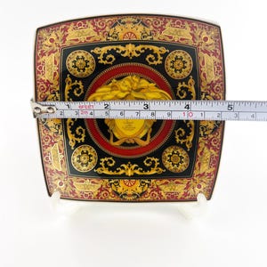 May include: Square decorative plate with a black, red, and gold ornate design. The plate features a central medallion with a yellow face and measures approximately 4 inches across.