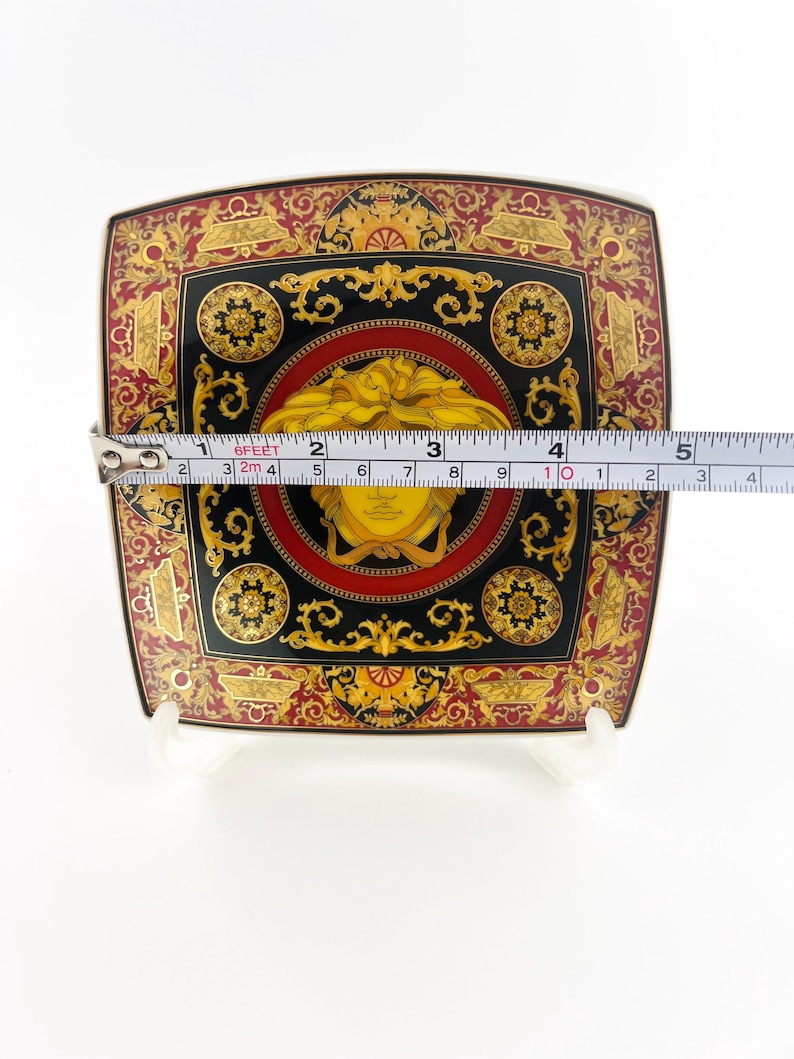May include: Square decorative plate with a black, red, and gold ornate design. The plate features a central medallion with a yellow face and measures approximately 4 inches across.