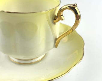 May include: A pale yellow teacup and saucer set. The cup and saucer have a scalloped edge and are trimmed with gold. The handle of the cup is also gold. The teacup is sitting on the saucer.