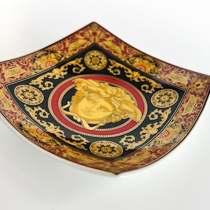 May include: A decorative square dish with a white base and a black, red, and gold ornate design. The center features a gold Medusa head within a red circle. The dish has a curved shape and a detailed border.