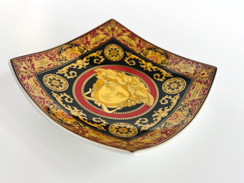 May include: A decorative square dish with a white base and a black, red, and gold ornate design. The center features a gold Medusa head within a red circle. The dish has a curved shape and a detailed border.