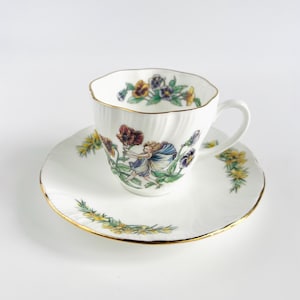 Può includere: Servizio da tè bianco con bordo dorato. La tazza e il piattino sono decorati con un motivo floreale e fatato nei toni del viola, giallo e rosso. La tazza ha un bordo ondulato.