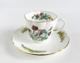 May include: White teacup and saucer set with a gold rim. The cup and saucer are decorated with a floral and fairy design in shades of purple, yellow, and red. The cup has a wavy edge.