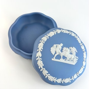 May include: A blue ceramic trinket box with a lid featuring a white relief design of figures and a chariot, surrounded by a white leaf garland. The box has a scalloped edge.
