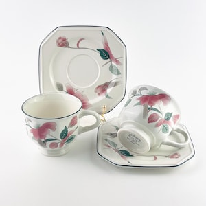 May include: A set of Mikasa tea cups and saucers. The white ceramic cups and saucers feature a floral design in pink, green, and gray. The edges of the cups and saucers are outlined in navy blue.