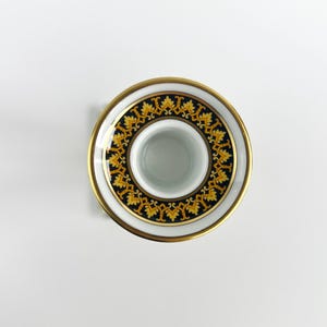 May include: A white ceramic candle holder with a gold rim and a decorative black and gold pattern. The design features a repeating motif of stylized leaves. The candle holder has a circular shape and a central opening for a candle.