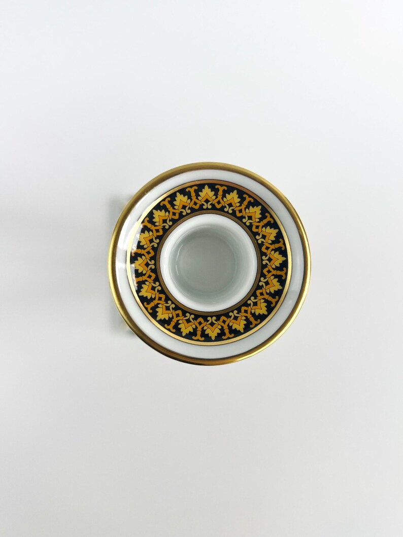 May include: A white ceramic candle holder with a gold rim and a decorative black and gold pattern. The design features a repeating motif of stylized leaves. The candle holder has a circular shape and a central opening for a candle.