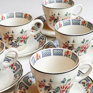 Puede incluir: Un juego de seis tazas y platillos blancos. Cada taza y platillo presenta un diseño floral y de pájaros con detalles en rosa, rojo y verde. Los bordes están adornados con un borde negro y un patrón geométrico azul y blanco.