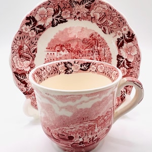 May include: A red and white teacup and saucer set. The cup and saucer feature a detailed landscape design with floral accents. The cup has a handle and the saucer has a scalloped edge. The set is made of ceramic.