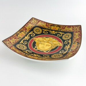 May include: Square decorative plate with a white rim. The plate features a black background with gold and red ornate designs. A gold Medusa head is centered on the plate, surrounded by a red circle.