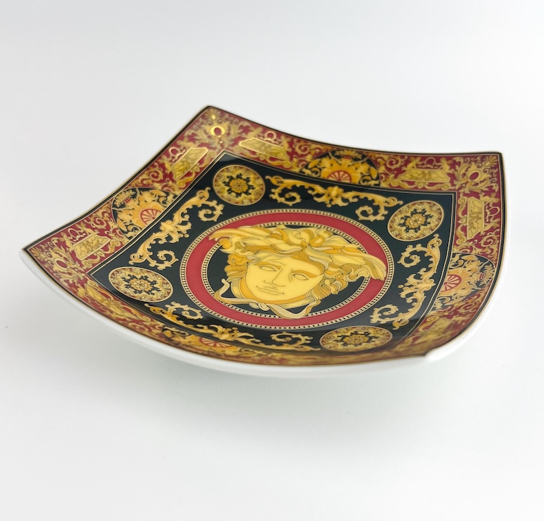 May include: Square decorative plate with a white rim. The plate features a black background with gold and red ornate designs. A gold Medusa head is centered on the plate, surrounded by a red circle.